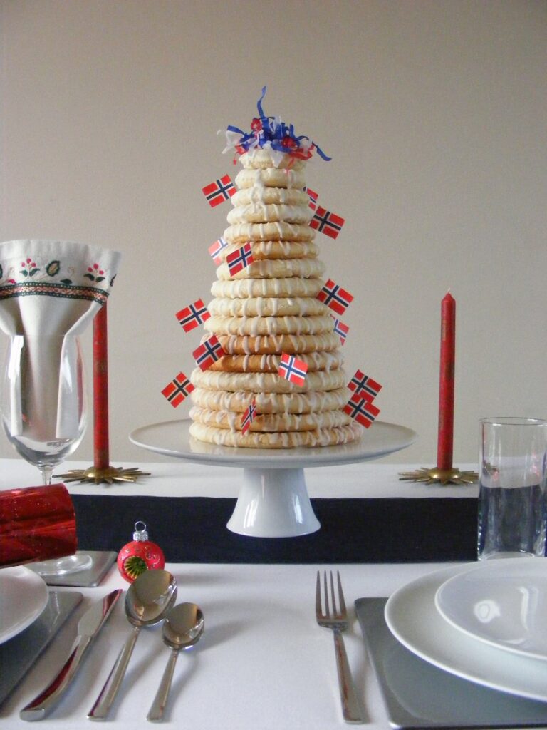 Image of a kransekake with sixteen increasingly smaller rings stacked on top of each other, drizzled with icing sugar and decorated with small Norwegian flags.