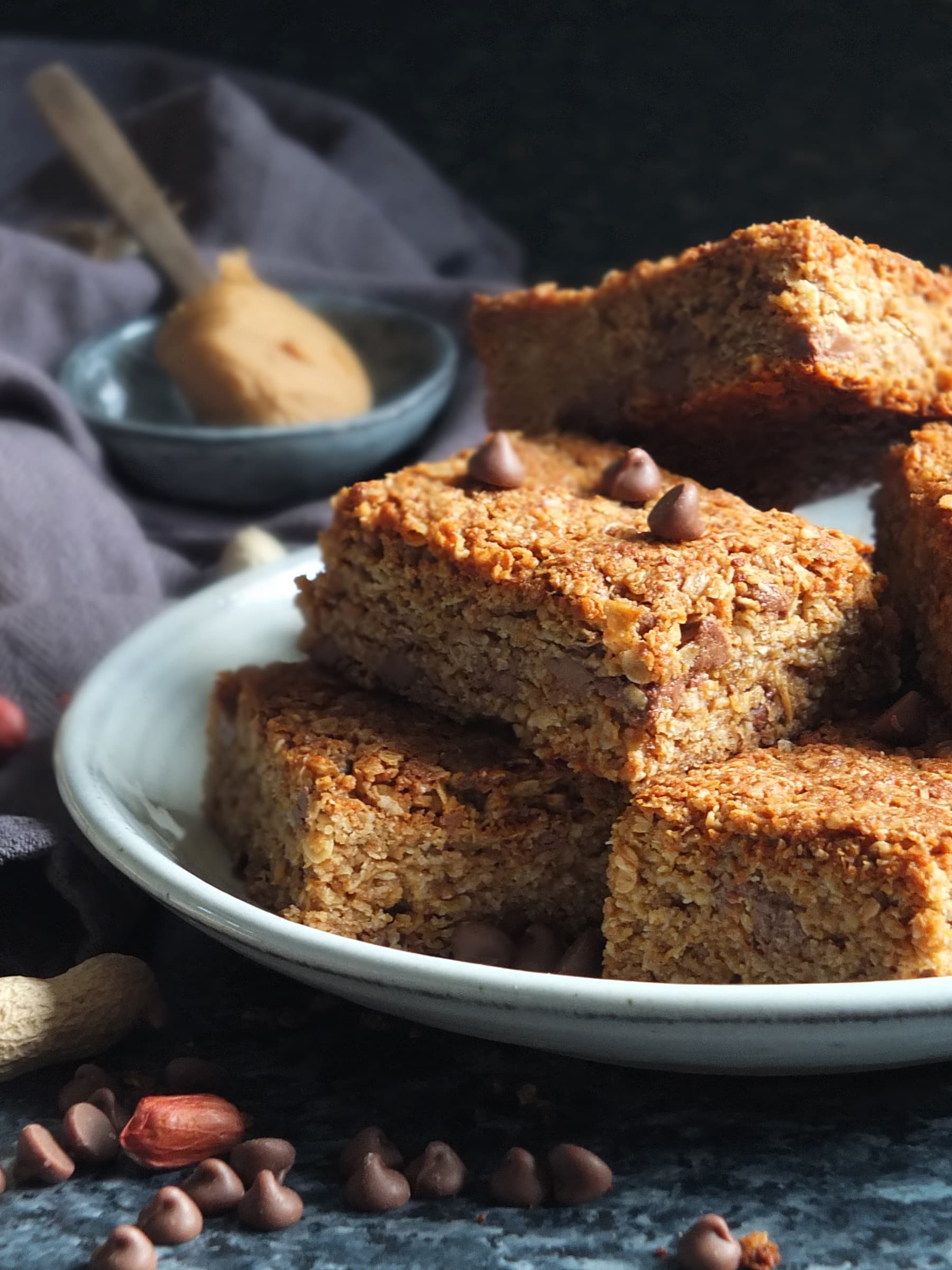 Moody and atmospheric image of peanut butter flapjacks on a serving plate in dark light, with chocolate chips and red peanuts decoratively laid out in the foreground, and a spoonful of creamy peanut butter in the background.