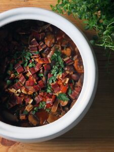 Top down image of a white pot filled with vegan mushroom bourguignon, garnished with crispy vegan bacon and chopped parsley.
